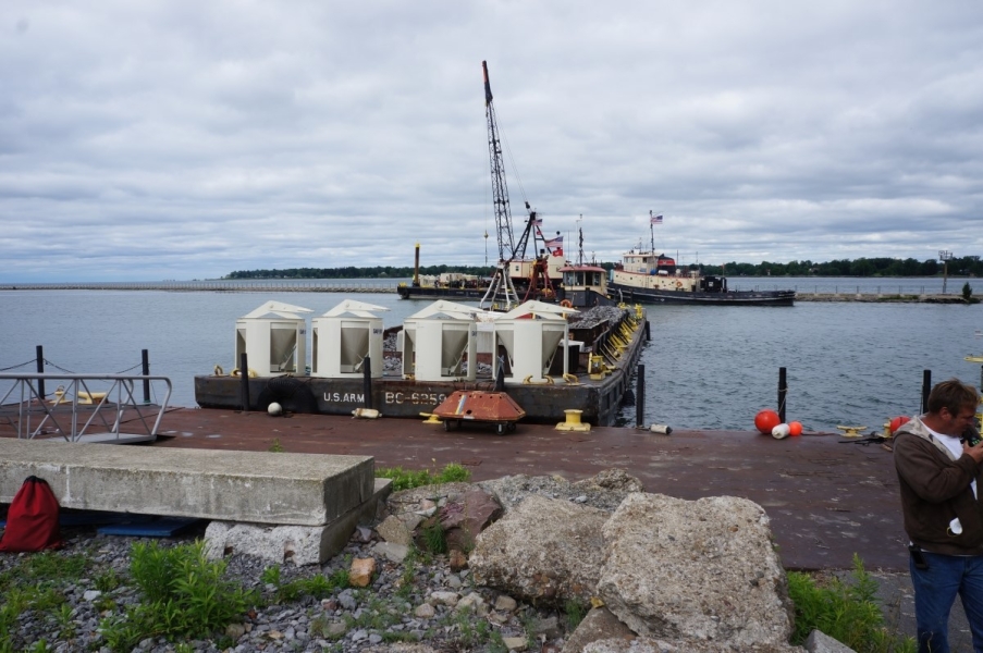 The front of a barge with some concrete equipment waits at a dock. There is a tug with a barge with a crane in the distance behind it.