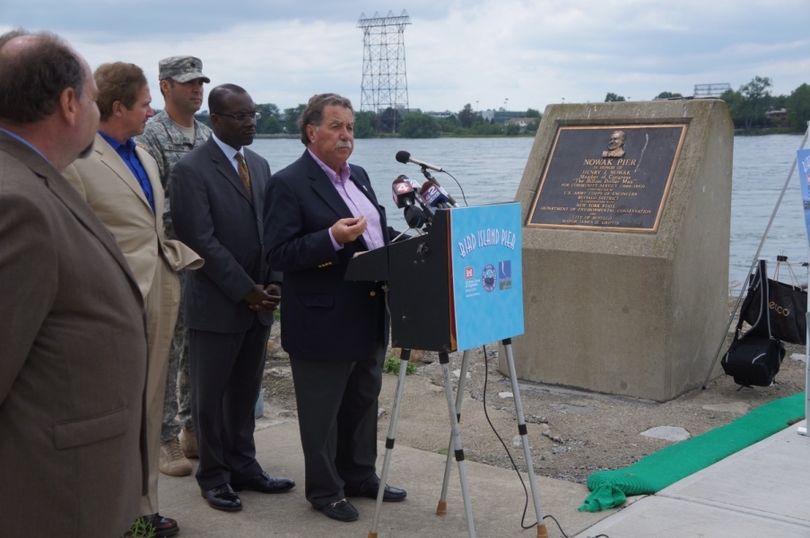 Five people in suits stand in front of a microphone at the waterfront. One person is wearing a military uniform. There is a plaque next to them.