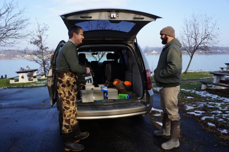 Two people stand at the open gate of a minivan with cinderblocks inside, in a parking lot by a river. There is some snow on the grass.