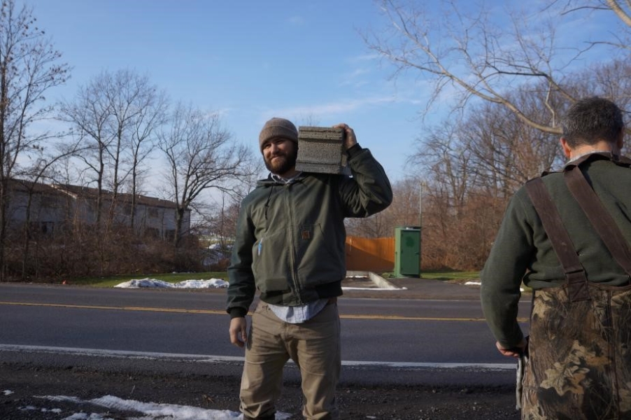 Two people by the side of a road, one holding a cinderblock on their shoulder.