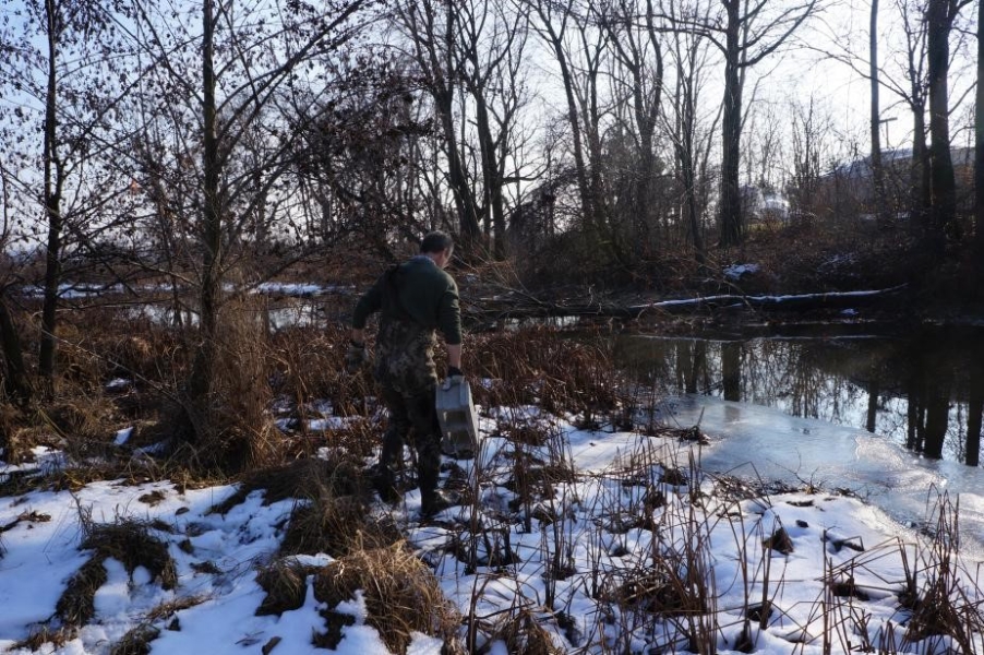 A person carrying a cinderblock walks on a snowy bank toward a creek.