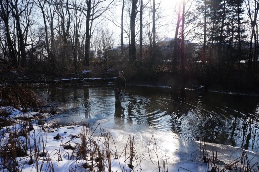 A person standing in the shallow water of an icy creek lowers a cinderblock into the water.