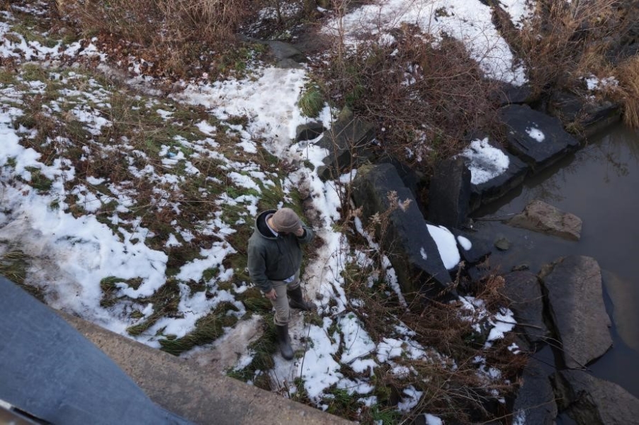 A person stands among rocks near the edge of water. There is snow on the ground. The picture is taken from above.