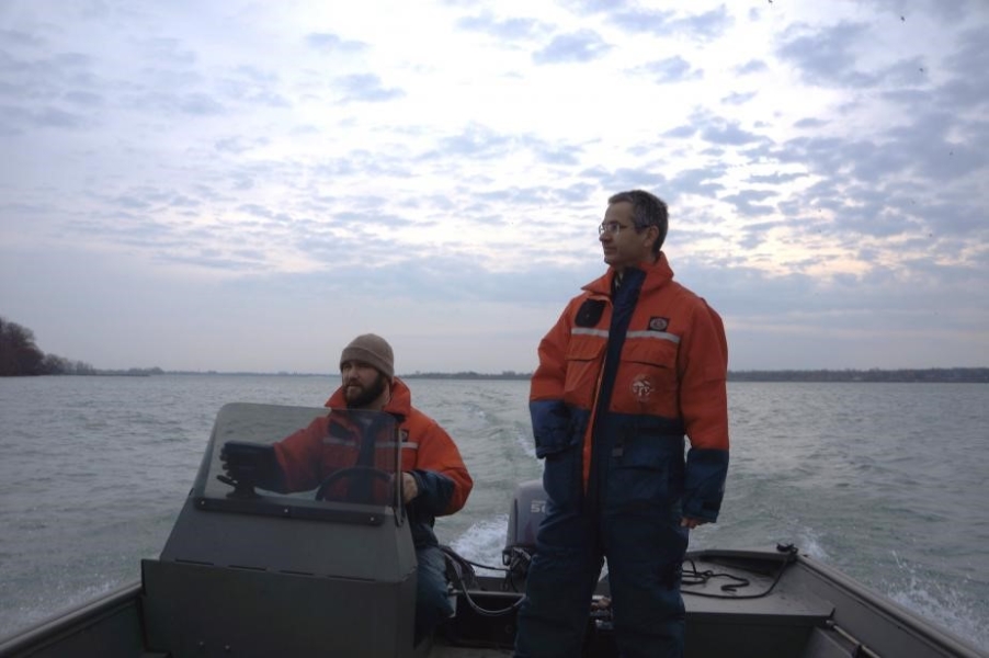 Two people in cold water safety suits on a boat. One is standing and the other sitting at the controls.