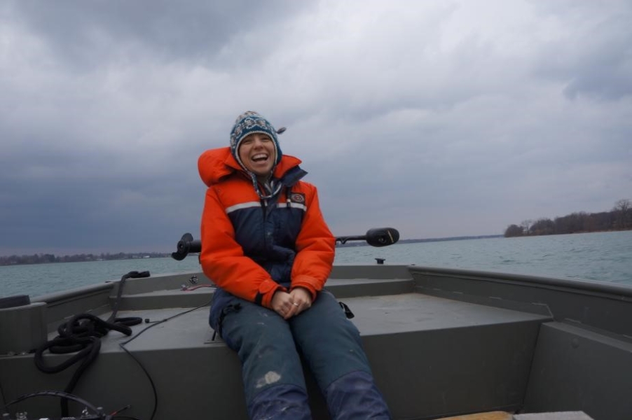 A person in a cold water safety suit and a knit cap smiles and sits at the front of a boat. The sky is cloudy.