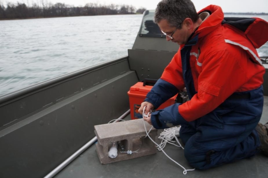 A person in a cold water safety suit kneels on a boat, tying a narrow rope around a cinderblock.