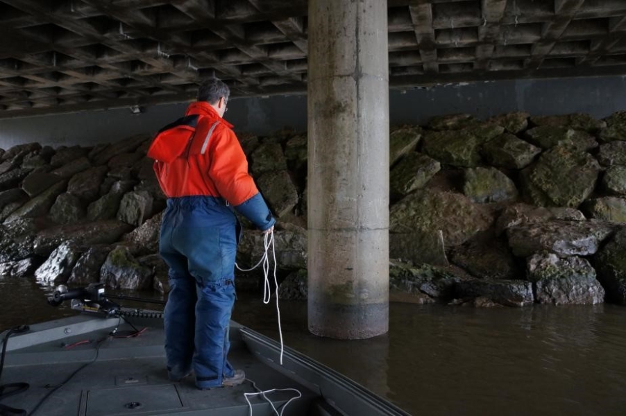 A person in a cold water safety suit stands on a boat under a bridge. There are rocks on the shore leading up to the bridge and a concrete pillar.