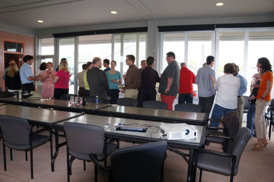 People standing and talking in a pavilion with tall glass windows all along one side. There is a table in the front of the picture with a projector set up on it.