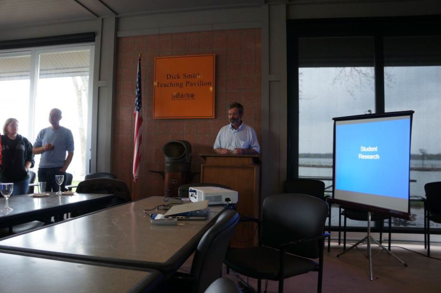 a person standing at the front of a room next to a small projector screen. Two people stand to the left.