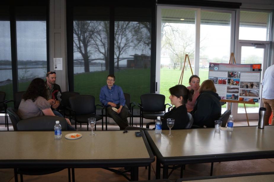 People seated and chatting, with a poster on an easel off to the side. There is a wall of windows behind them.