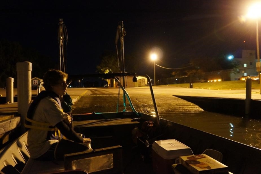 A person in a life jacket sits on a boat at a boat launch at night