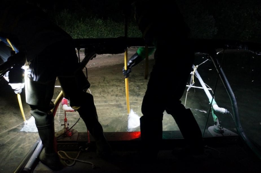 Nighttime picture of two people standing at the edge of a boat scooping nets on long poles through the water.