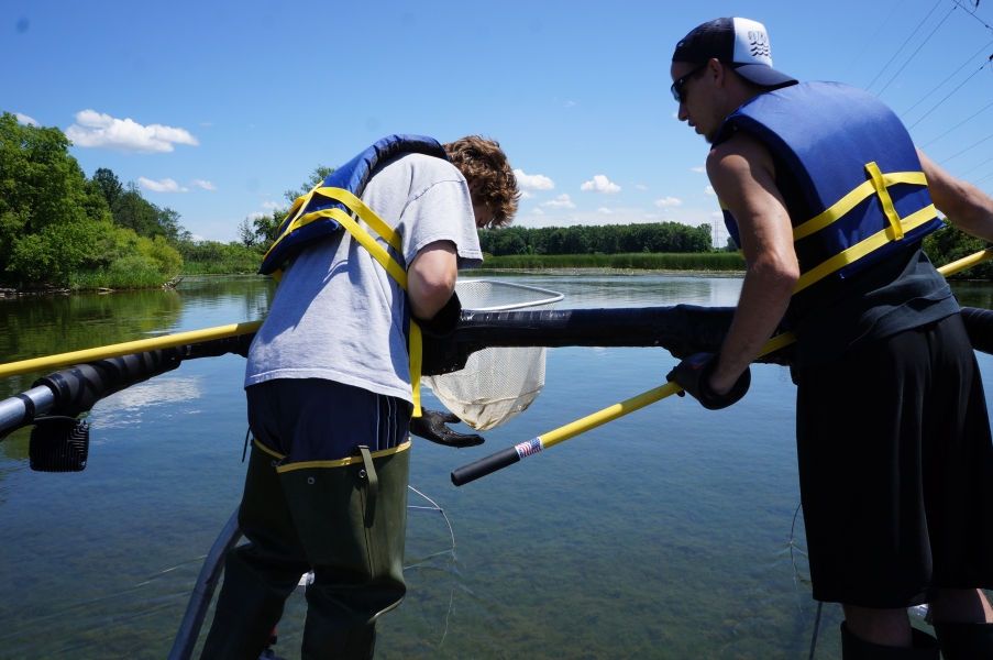 Two people stand at the front of a boat holding nets on long poles. One is looking at something in their net, while the second glances over at the net too.