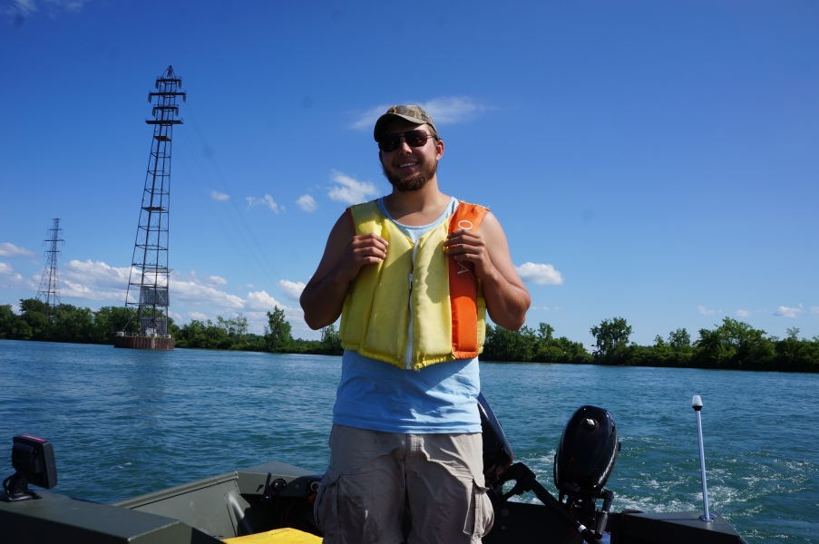 A person in a life jacket smiles and stands at the back of a boat on a river. There are high-power electric towers in the background.