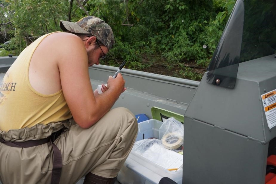 a person wearing chest waders kneels on a boat and writes information on a bottle