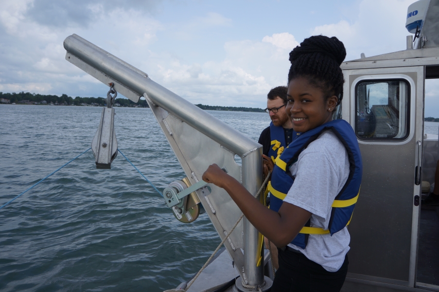 two people in life jackets stand on a boat. One smiles and turns the crank of a winch while the second watches the equipment in the water