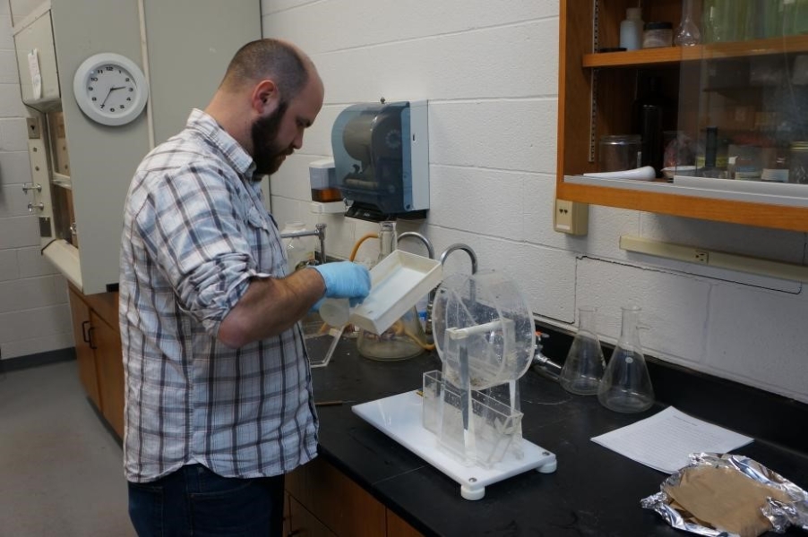 a person standing at a counter in a lab wears gloves and holds a small white pan. There is a clear Plexiglas wheel sitting on the counter.