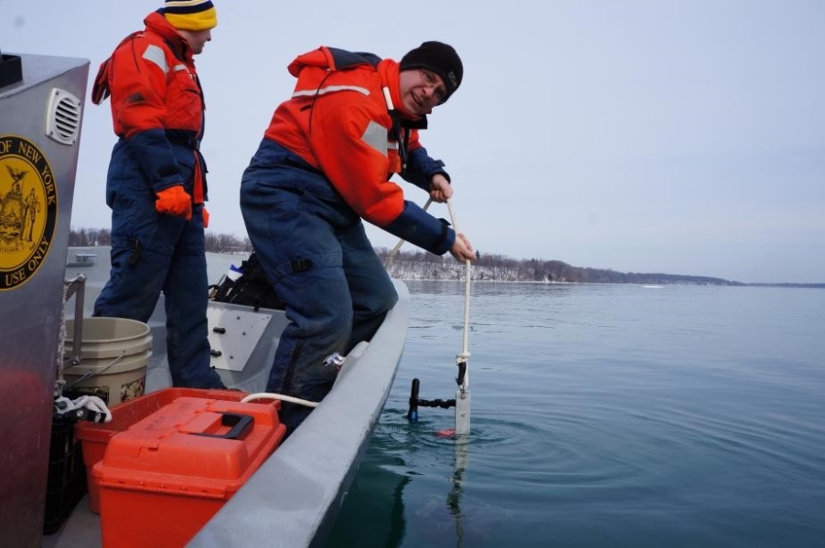 Two people in coldwater safety suits stand on a boat. One person lowers something over the side of the boat