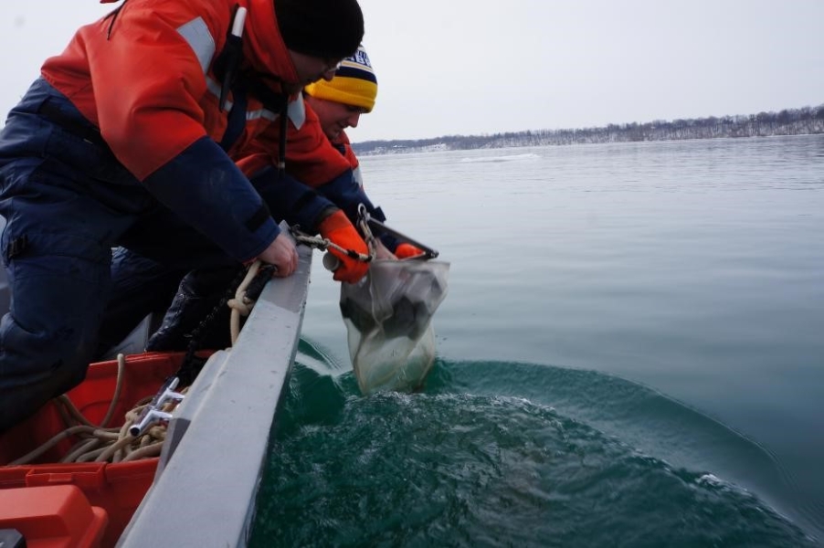 Two people in coldwater safety suits work to bring in sampling equipment over the side of a boat