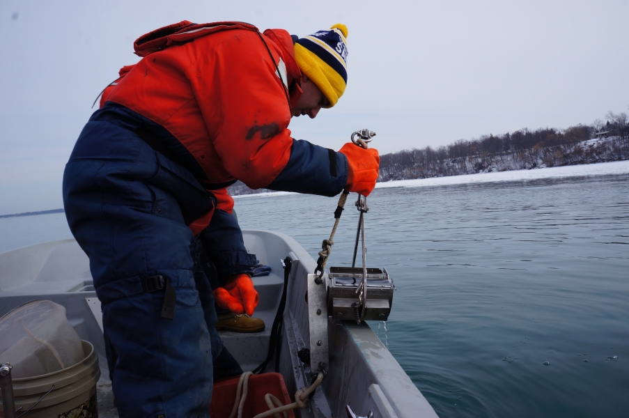 a person in a flotation suit, hat, and gloves holds a metal contraption on the edge of a boat in an icy river