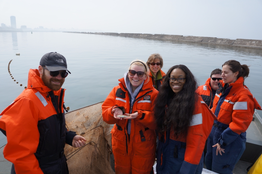 People in cold water safety gear stand on a boat near a breakwall. One person holds a large square-hooped net, while another holds a small fish on their hands.