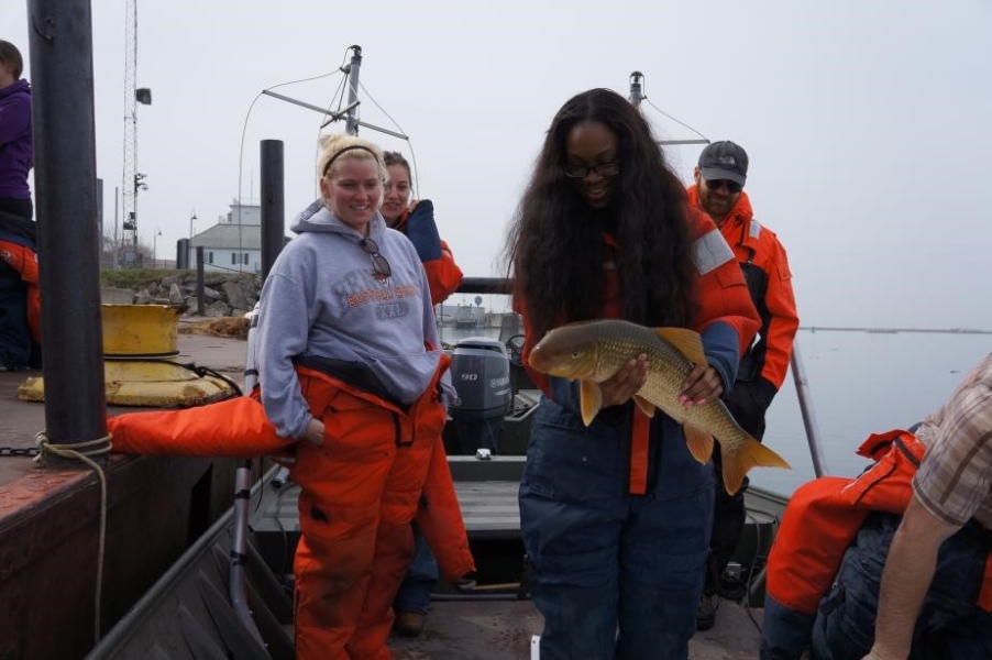 A few people stand on a boat at dock. One holds up a two-foot long brown and white fish with orange fins