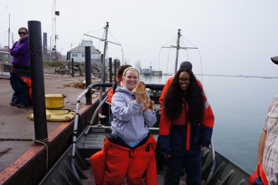 A few people stand on a boat at dock. A different student holds up a fish