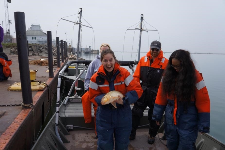 A few people stand on a boat at dock. A third different student holds up a mostly white fish