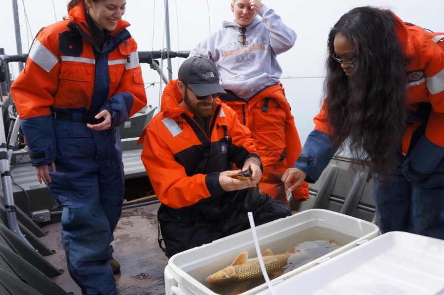 Students stand around a cooler with water and at least one large fish. A student in the center holds a small brown fish in their hands