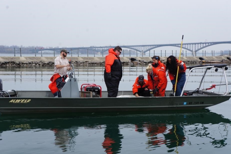 Students on a shallow boat with railings and two booms. Some of them are looking at a cooler