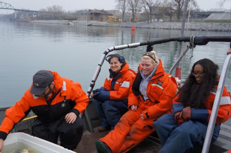 Students sit at the front of a boat near the railings, while the student at left crouches over a cooler with water and fish in it.