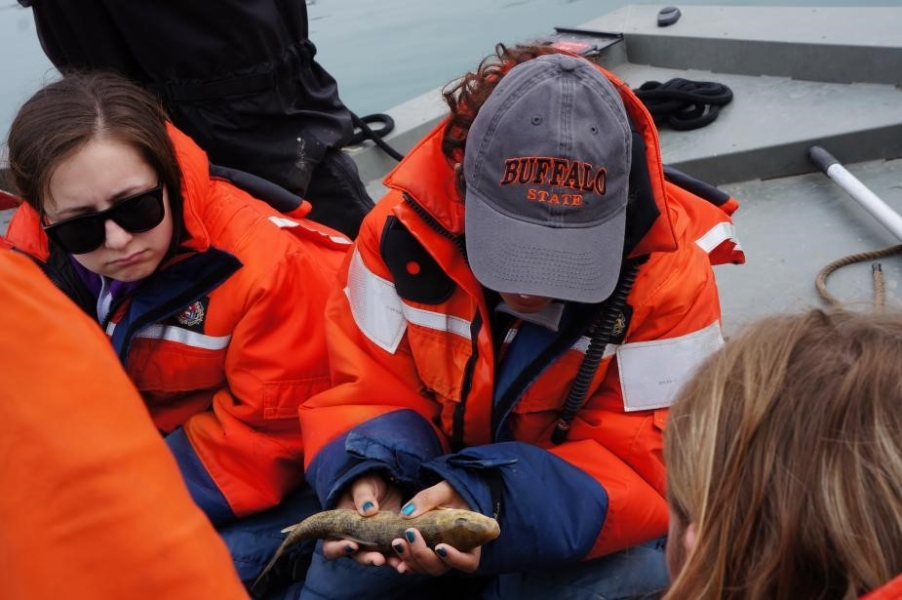 A few people sit around a person with a Buffalo State hat who is holding a medium-sized brownish fish