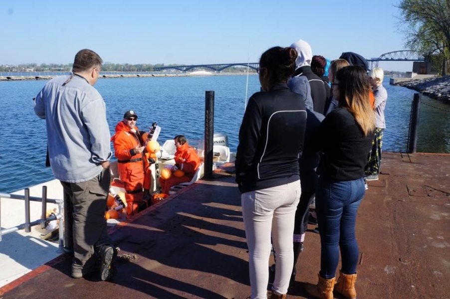 Several people stand on a dock by a boat that is moored there. There is a person in the boat holding up an instrument with a flotation device on it.