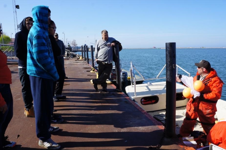 Several people stand on a dock by a boat that is moored there. There is a person in the boat holding up an instrument with a flotation device on it.