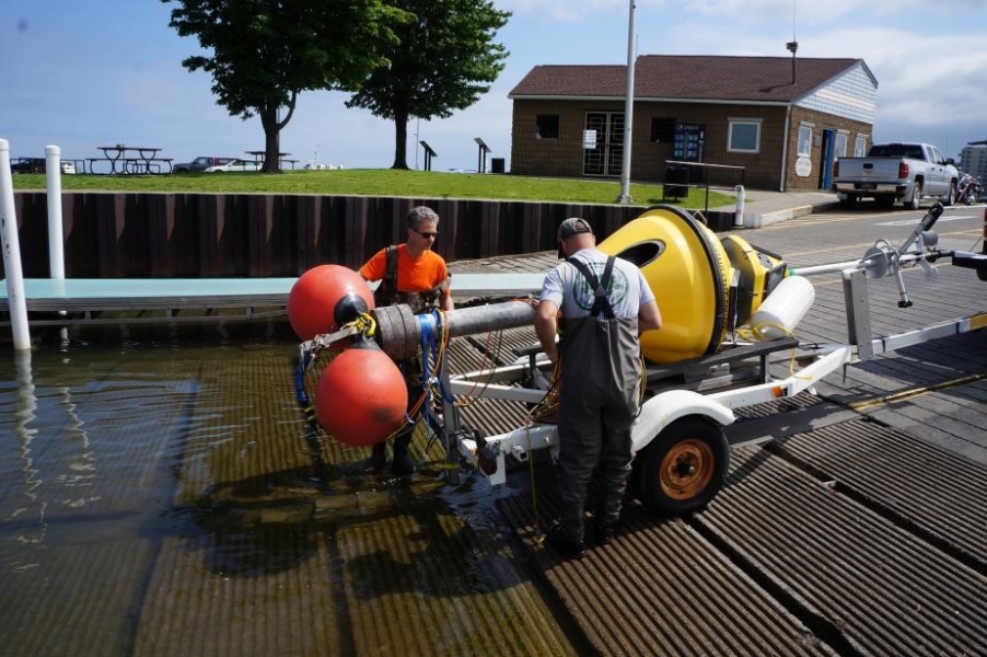 Two people stand next to a buoy strapped to a boat trailer. The buoy has been backed down a boat ramp and there are floats tied to the buoy.