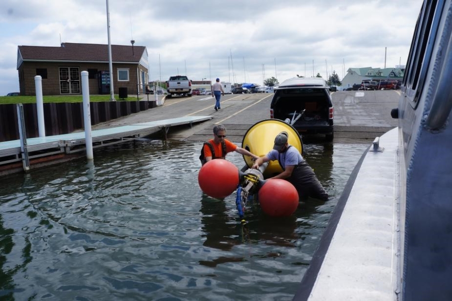 Two people wearing chest waders stand in shallow water at a boat launch to maneuver a buoy on a trailer. There are two floats tied to one end of the buoy.