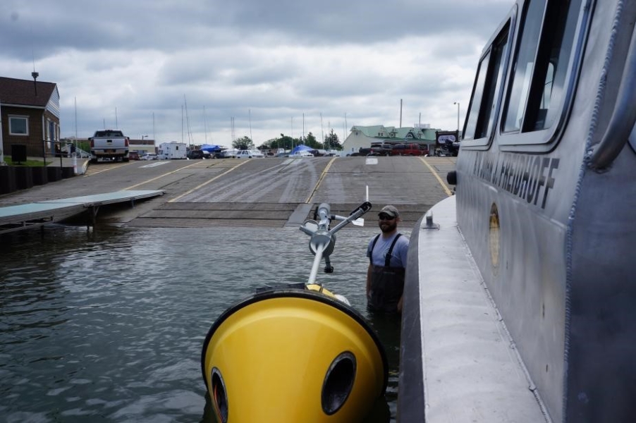 A person in chest waders stands in shallow water next to a buoy floating on its side next to a boat at a boat launch.