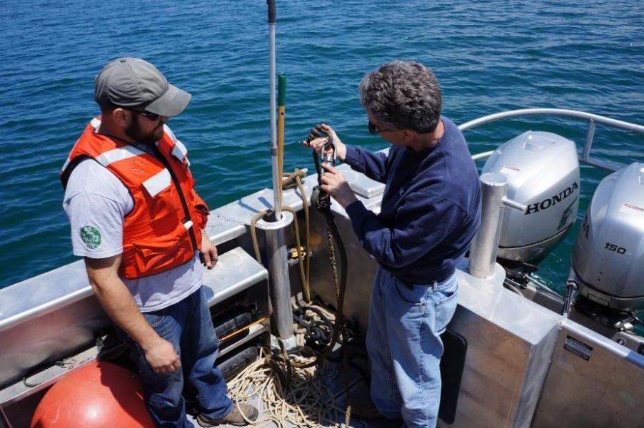 Two people stand on the back of a boat. One is holding up a line with shackles on it to connect two sections of rope together.