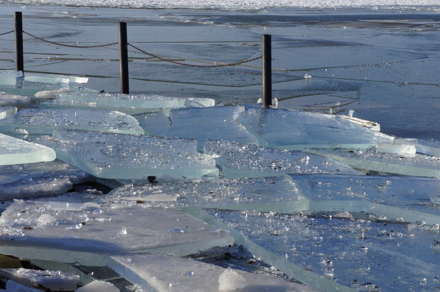 Broken sheets of ice on a surface by the water. Light sparkles in smaller pieces of ice. The sheets of ice are at least 4 inches thick.