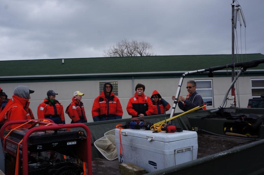 Students in flotation suits stand behind a boat with a cooler and a generator on it. There is a single story building behind them.