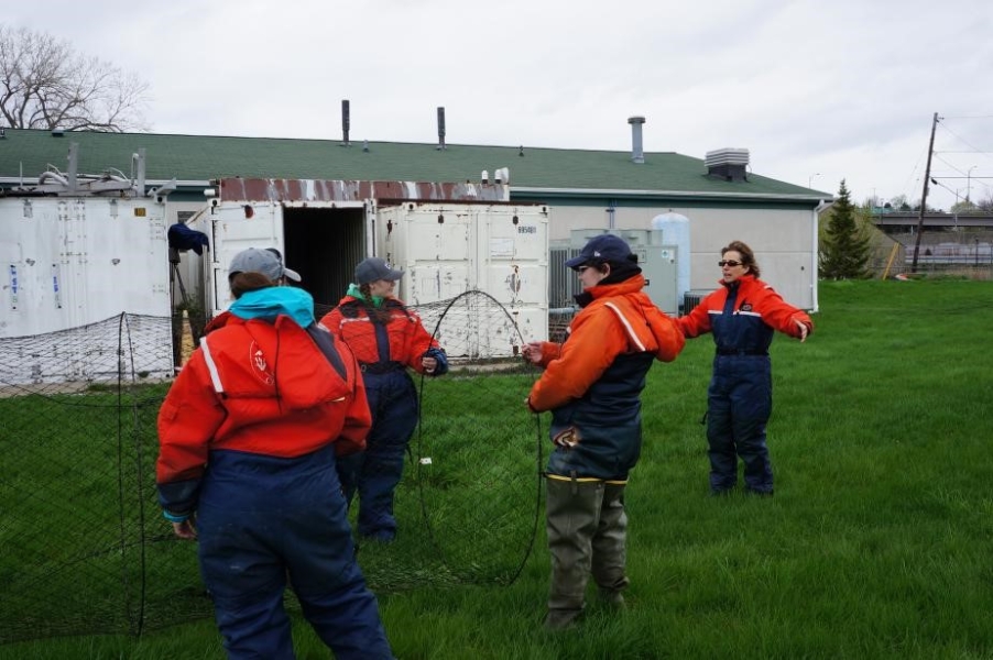 Three students in flotation suits hold a conical trap net open, while a fourth person gestures behind them, during an on-land demonstration. There is a building in the background.