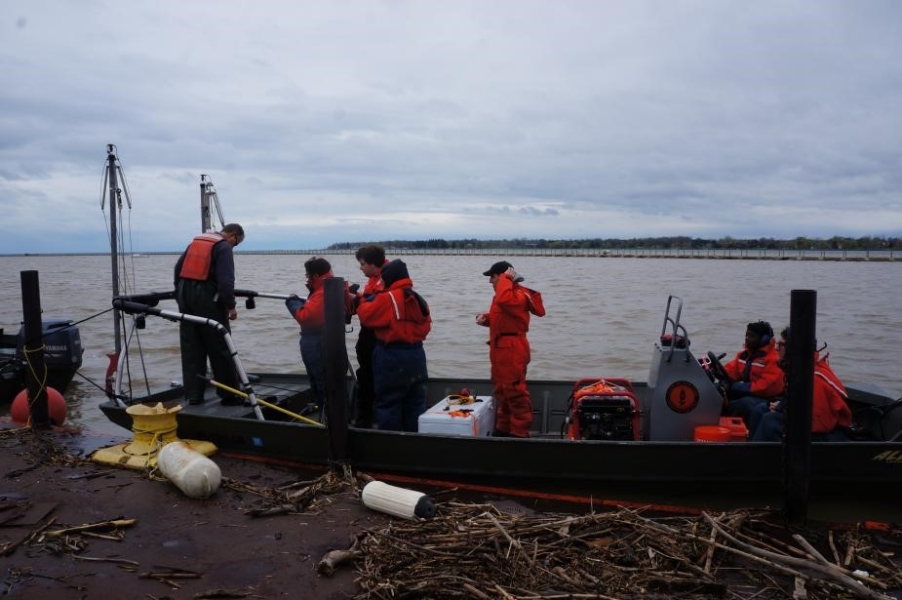 People in flotation suits stand on a flat boat tied up at dock. There is woody debris on the dock nearby.