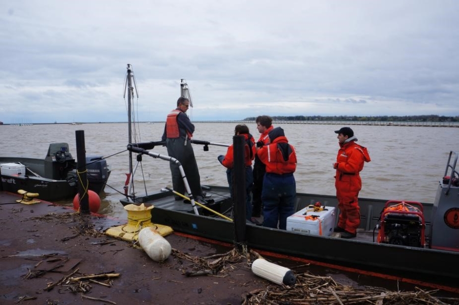 People in flotation suits stand on a flat boat tied up at dock. There is woody debris on the dock nearby. A second boat is tied up nearby.