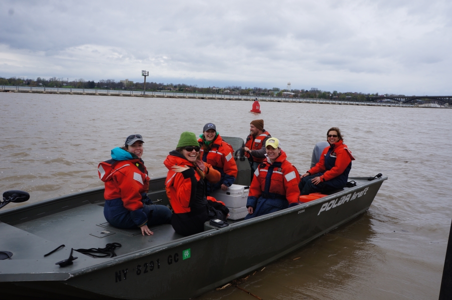 Six people in coldwater safety gear on a small boat in a muddy river on a cold day. Most are smiling or waving at the camera while one drives