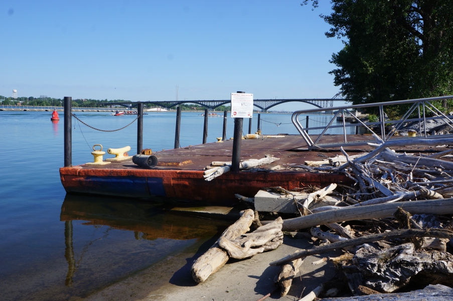 A dock made from a rusty sunken barge. Piles of driftwood sit on the concrete next to the dock.