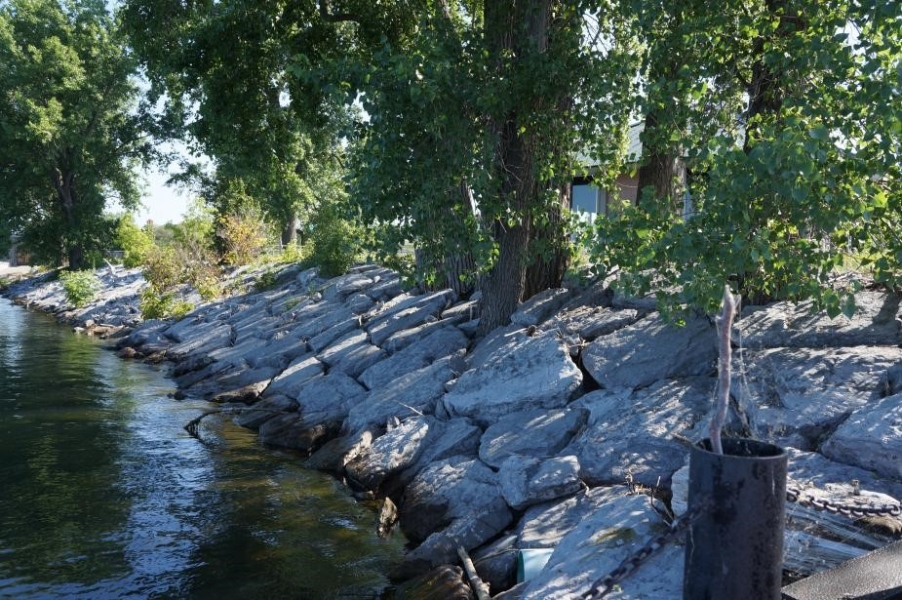 Neatly stacked stone slabs on the slope between the ground and the water, under a tree.