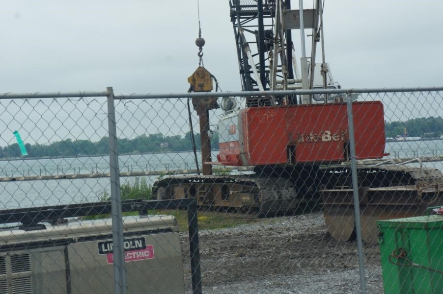 A crane with a jackhammer device on the end of its cable, driving a metal girder into the water in front of it.