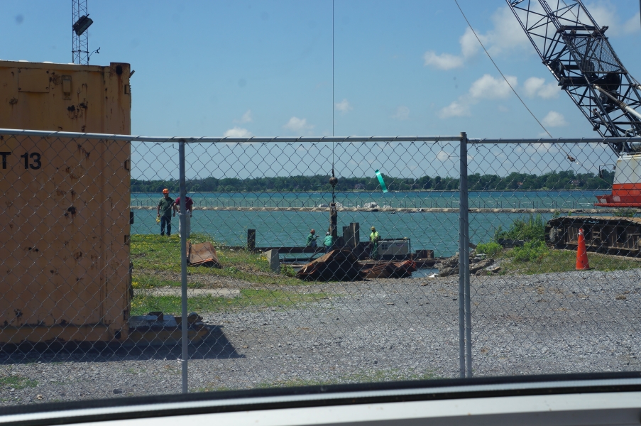 View of a construction site at the waterfront through a fence. A crane is lowering a steel beam down to some workers.