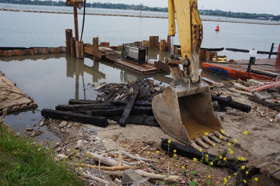 A construction zone at the edge of the water. There is a wall of steel surrounding the end of a boat ramp, which is full of water. At the end of the ramp are some blackened beams of wood. The bucket of an excavator rests on the ramp.