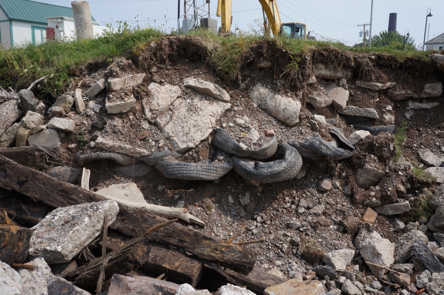 The side of excavated ground showing layers of dirt, stone, rubber tires, and wooden beams.